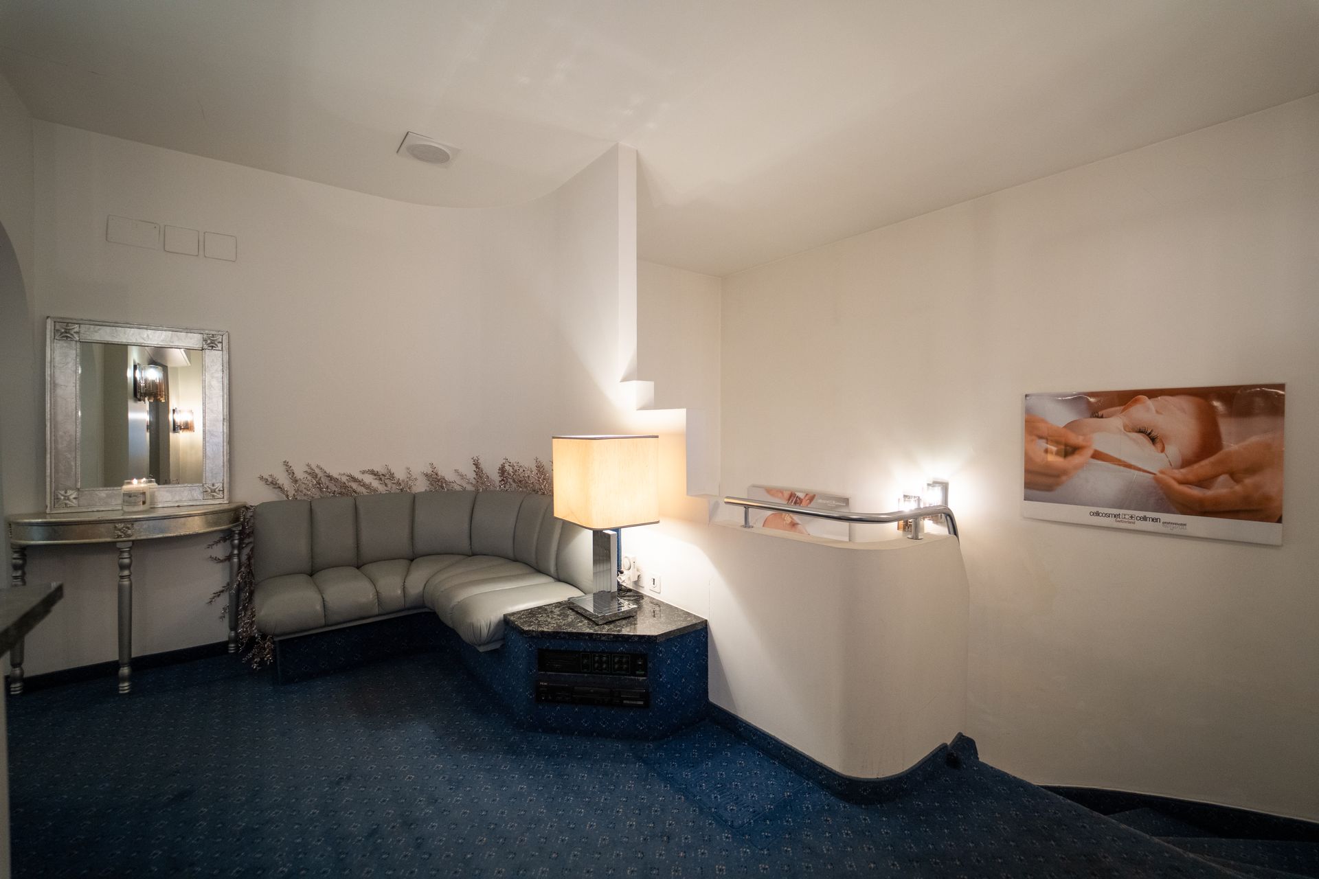 Wartebereich mit grauem Sofa, blauem Teppich, Lampe, Schminkspiegel und Wandbildern, die eine Spa-Behandlung darstellen.