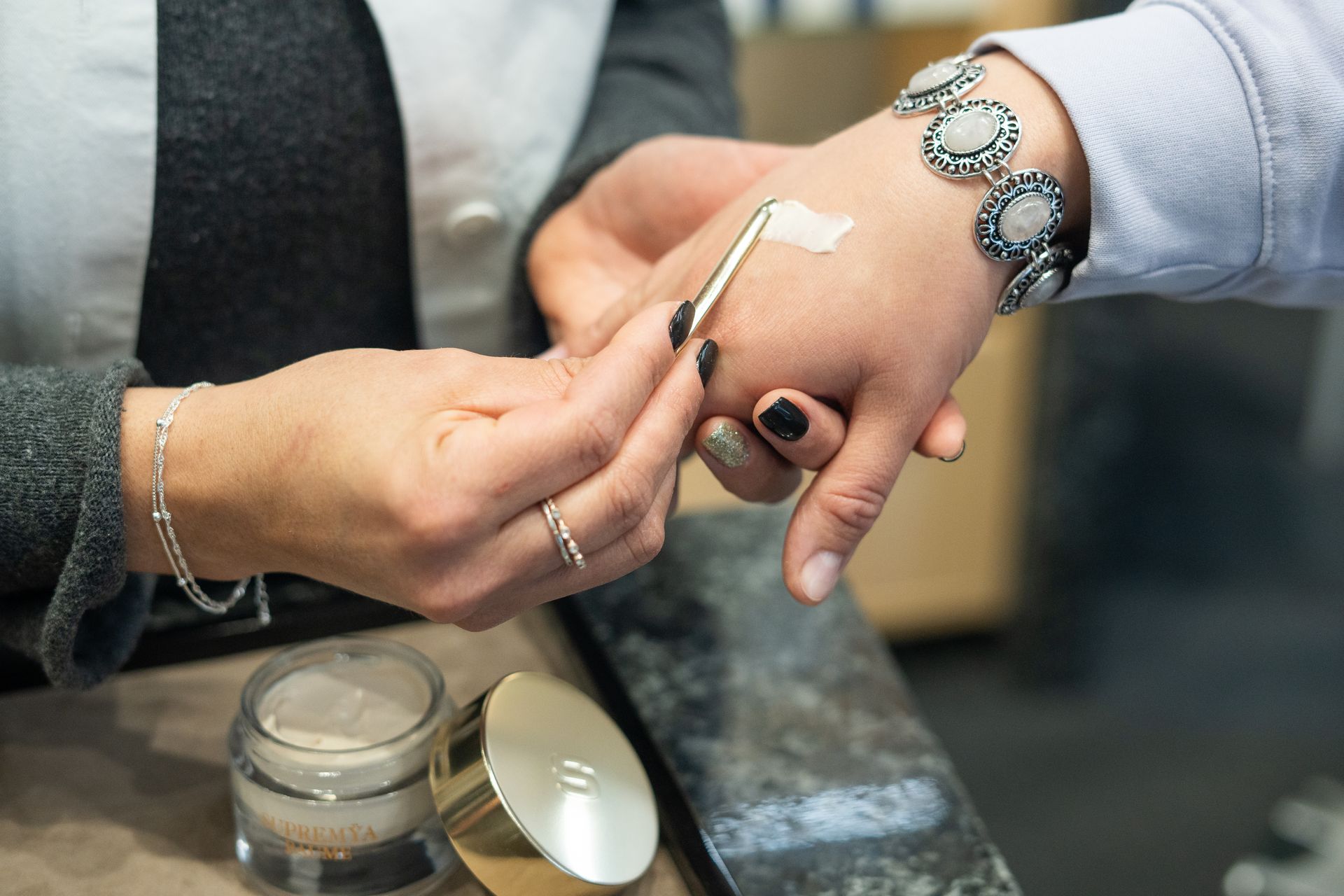 Kosmetikfachkraft trägt mit Spatel helle Creme aus Tiegel auf Hand einer Kundin auf, professionelle Beratung am Stand.