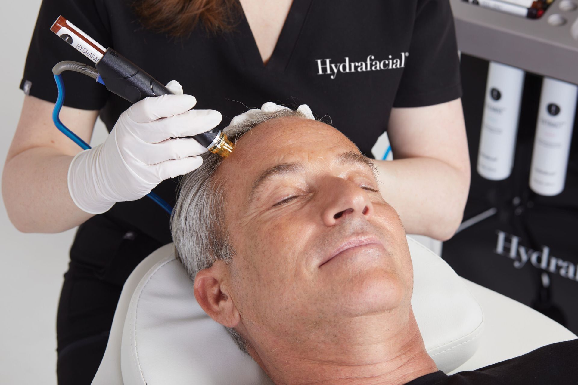Eine Person, die sich von einem Behandler in einem schwarzen Marken-Top einer Hydrafacial-Kopfhautbehandlung unterzieht.