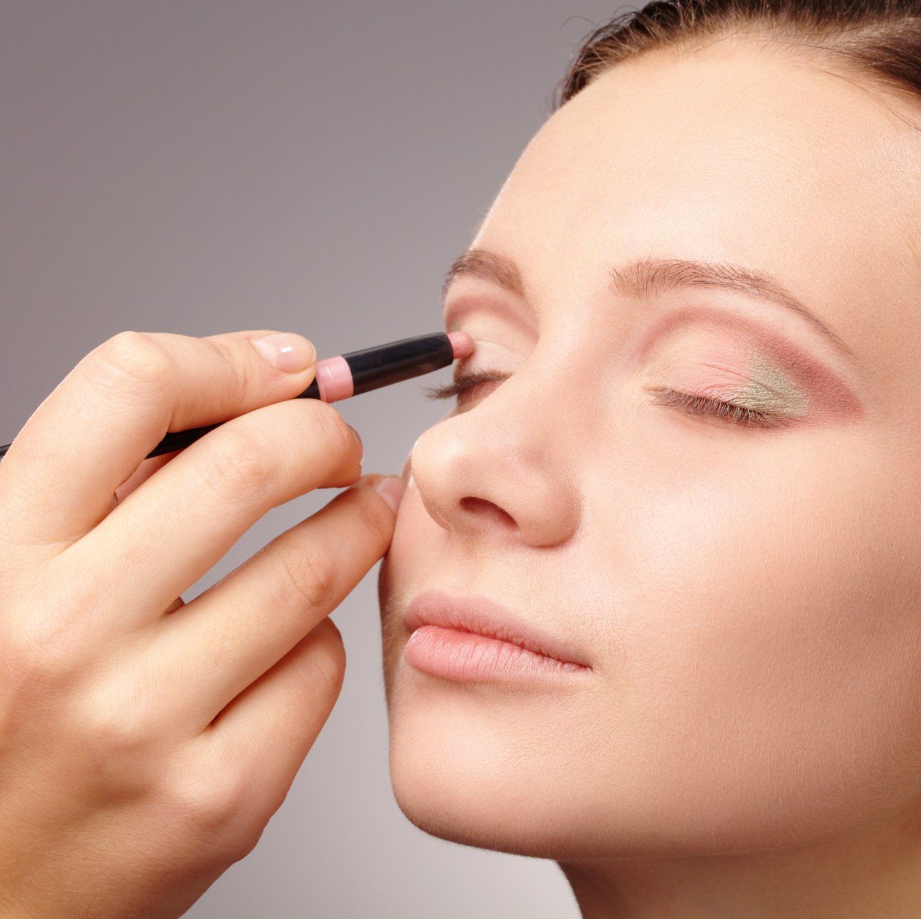 Nahaufnahme einer Hand, die mit einem Make-up-Stift einen zartrosa Cremelidschatten auf das Augenlid aufträgt.