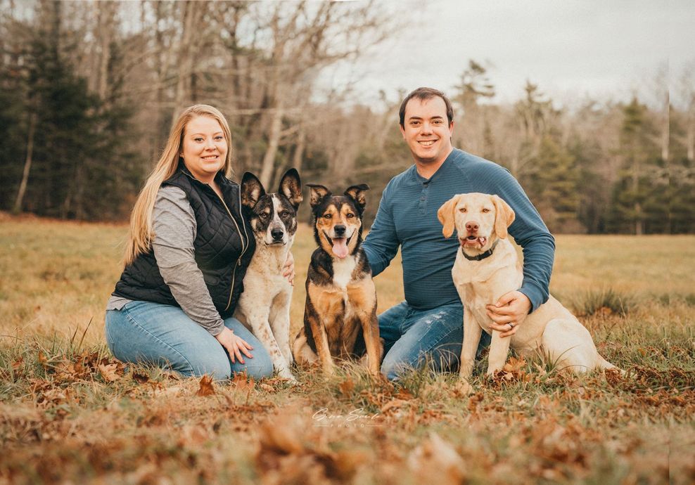 Couple kneels with three dogs in a field. Woman smiles with black vest, man smiles in blue shirt.
