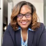 A woman wearing glasses and a blue shirt is smiling for the camera.