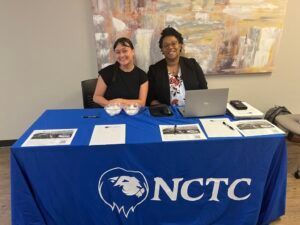 Two women are sitting at a blue table with a laptop.