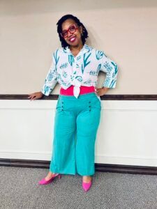 A woman wearing a white shirt and green pants is standing next to a wall.