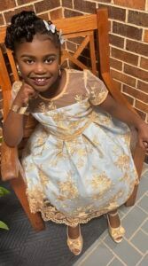 A little girl in a white and gold dress is sitting in a chair.