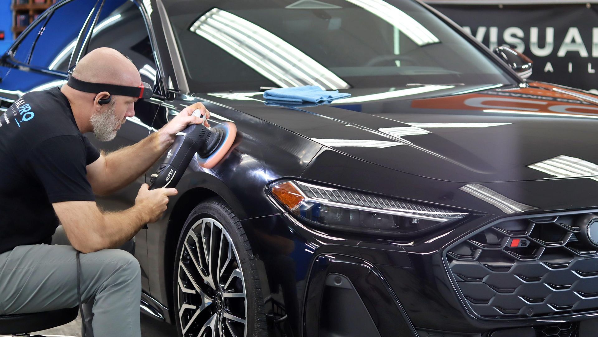 Technician polishing a dark blue sports car in a garage, focusing on the front fender and headlight.