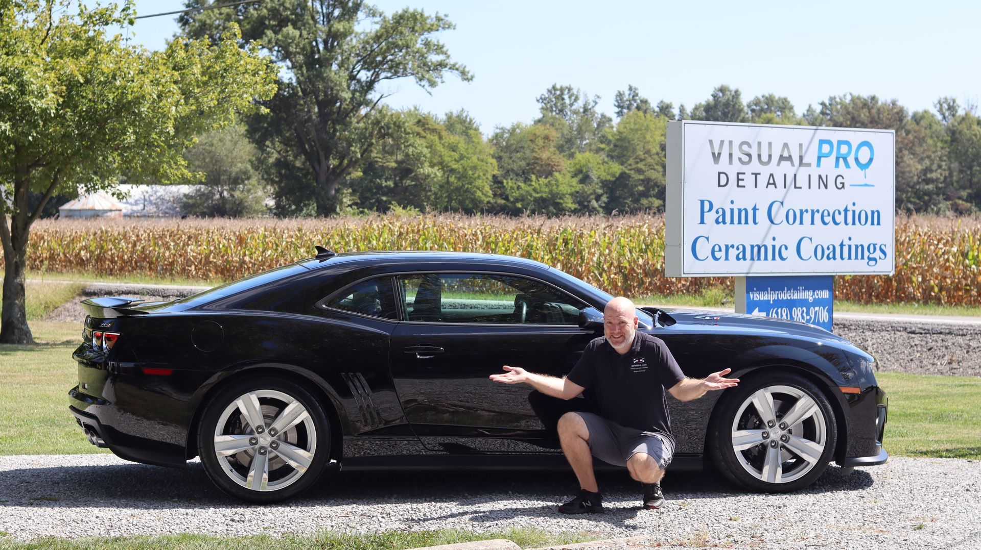 Black Camaro parked in front of a sign for a detailing business; man crouches beside it, gesturing with hands.