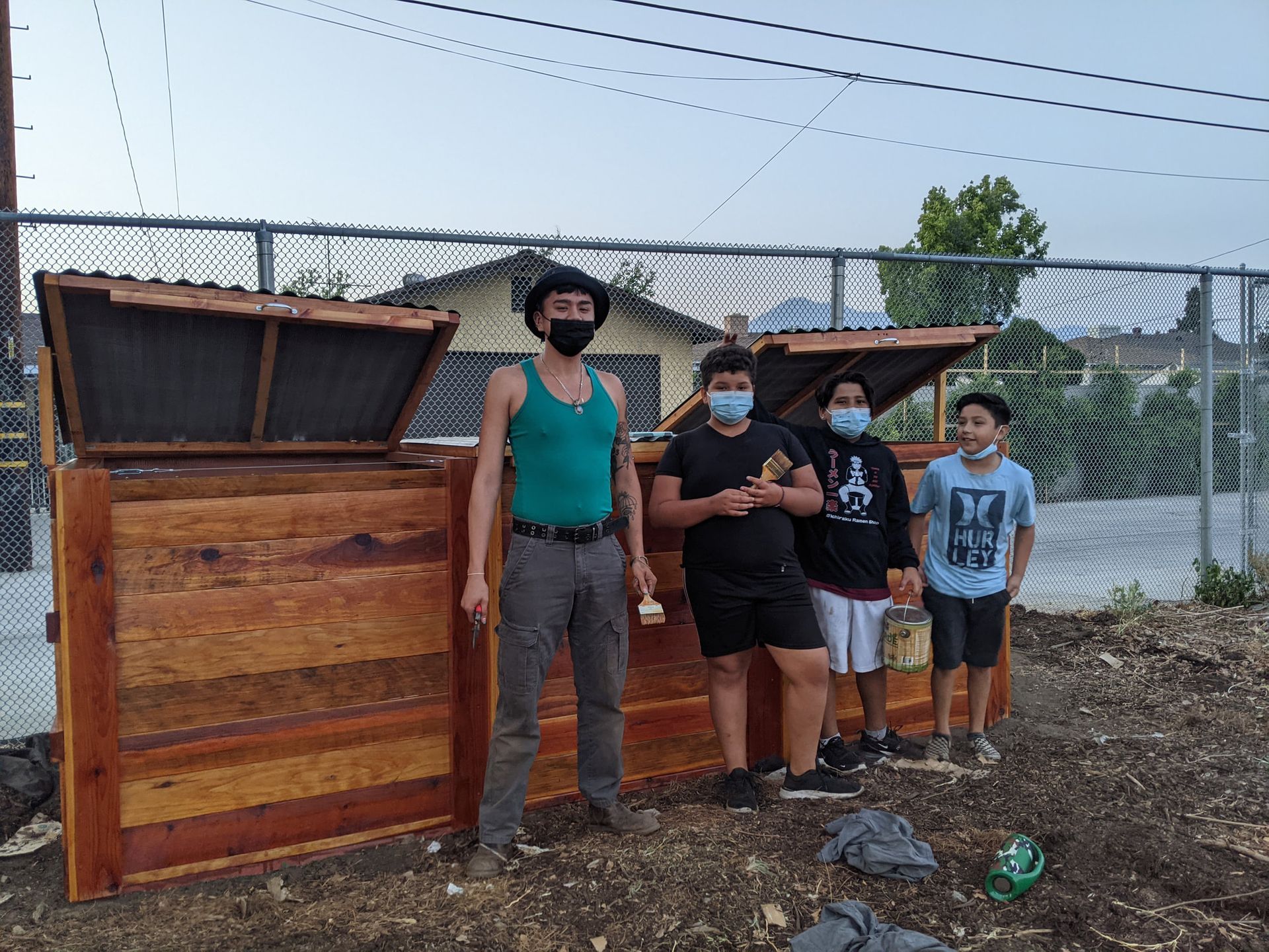 Four people stand in front of two newly built wooden compost bins outdoors near a chain-link fence.