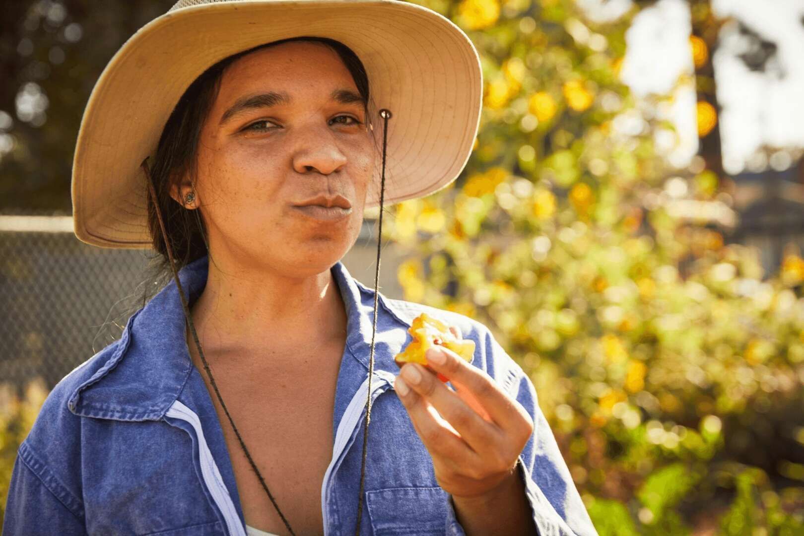 A person wearing a wide-brimmed sun hat and blue shirt tastes a piece of fruit in a sunny outdoor garden setting.