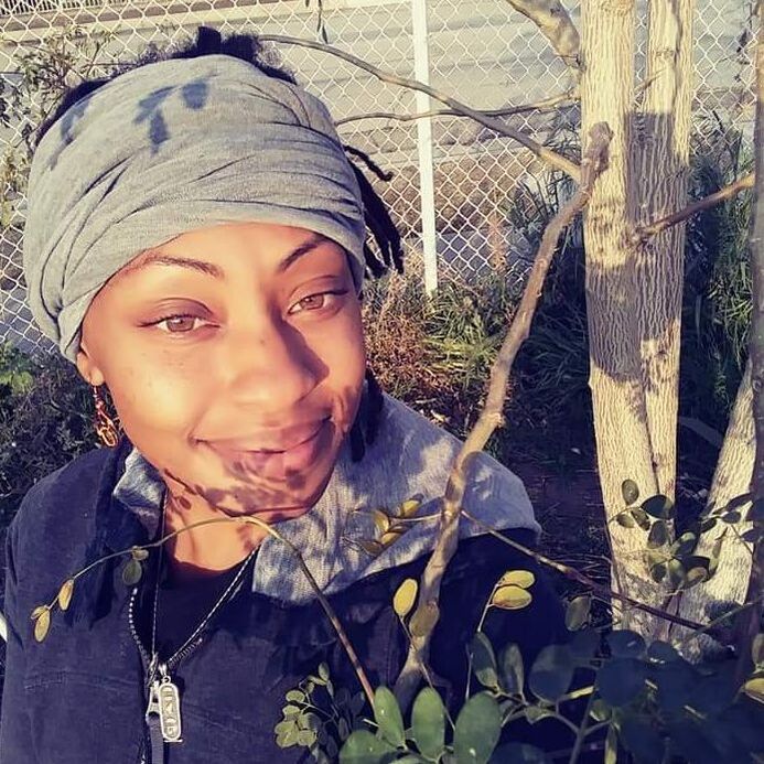 A person with dark hair and a gray headwrap smiles outdoors, with sunlight casting tree branch shadows on their face.