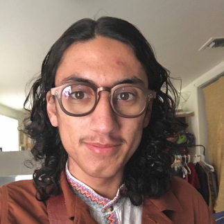 A person with long, dark, wavy hair and glasses smiling softly, wearing a brown jacket over a patterned collared shirt.
