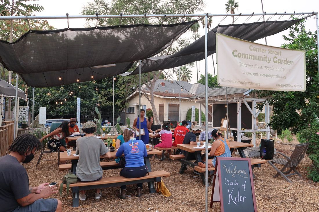 People sit at wooden tables under a shade canopy at the Garden Hills Community Garden.