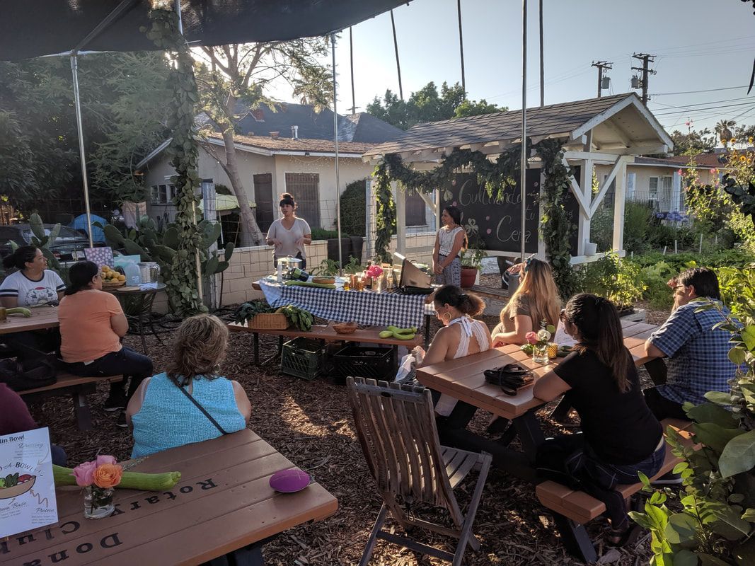 People sit at outdoor tables in a garden setting, listening to a speaker standing near a decorated trellis and a house.