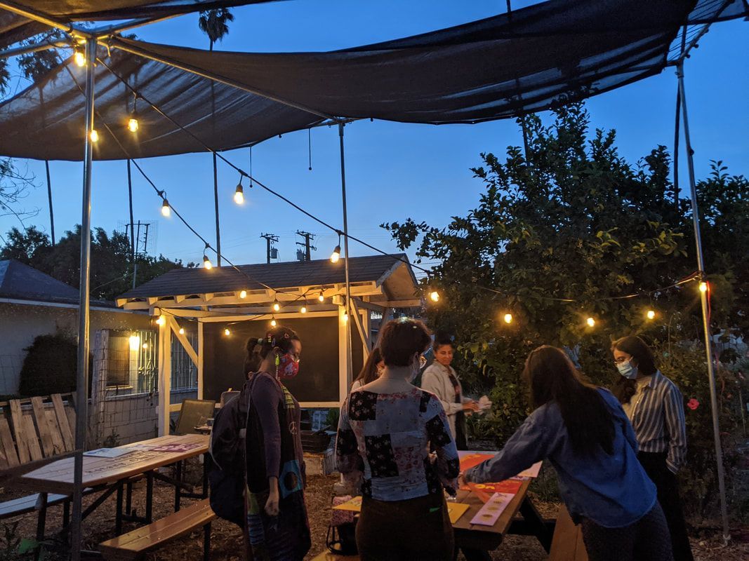 People stand around tables under string lights and a shade canopy in an outdoor area at dusk.
