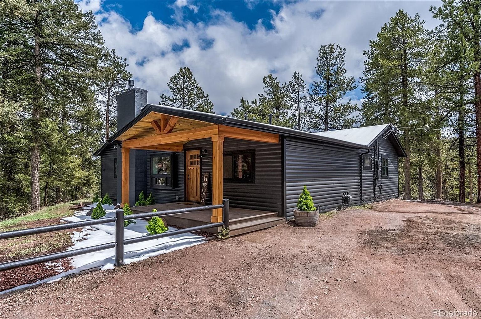 A small black house with a wooden porch is surrounded by trees.