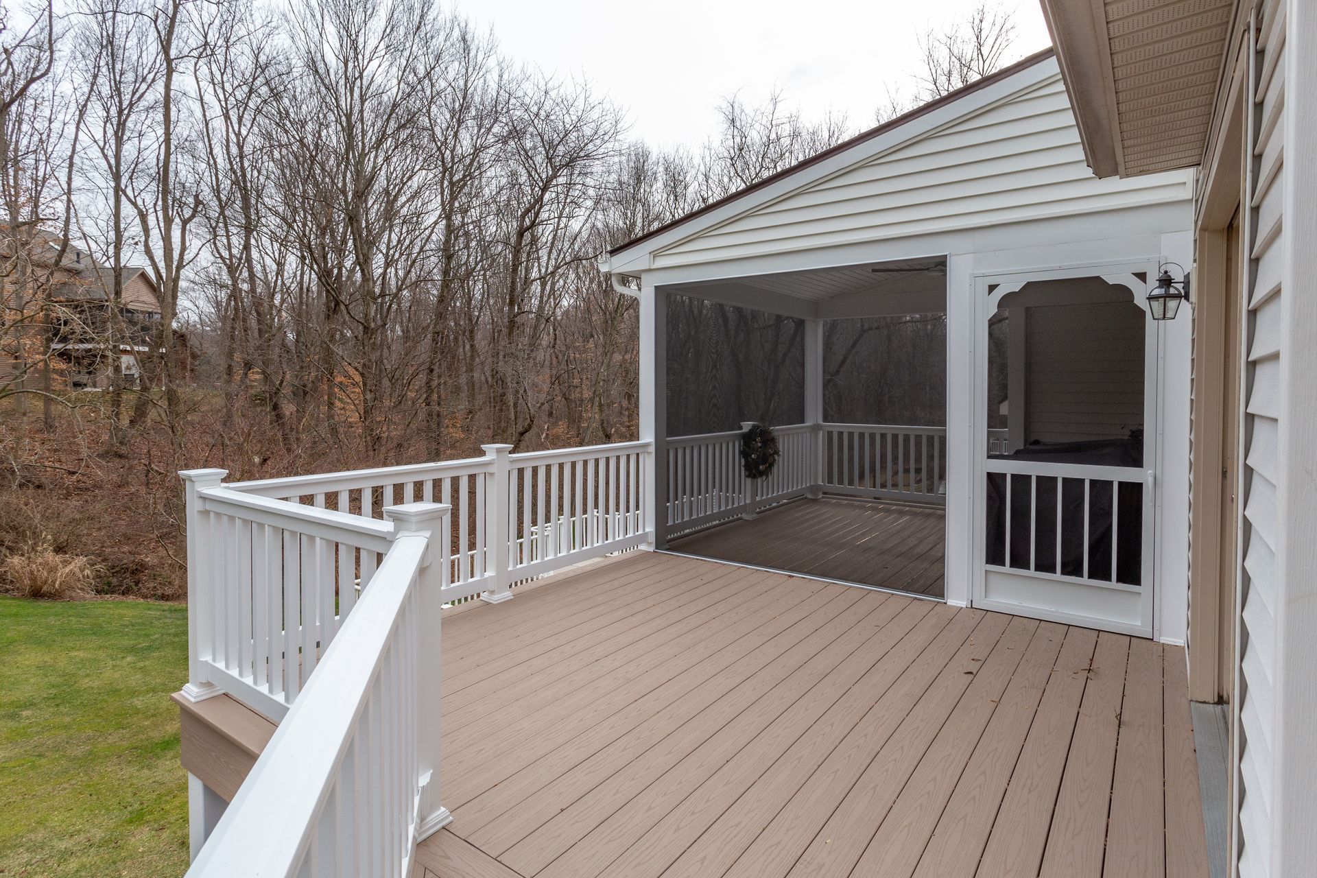 Covered wooden deck with white railings beside a house, overlooking bare trees and a wooded yard