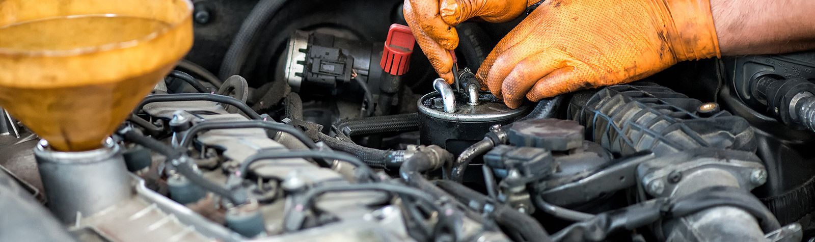 Hands in orange gloves working on a car engine, with a yellow funnel visible.