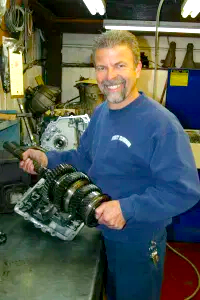 A man in a blue sweatshirt is holding a gearbox in a garage.