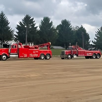 Two red tow trucks parked on a dirt lot, with trees and a cloudy sky in the background.
