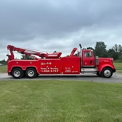 Red tow truck parked on grass, with an extended boom under an overcast sky.