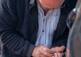 Man in glasses working on something outdoors, wearing a dark jacket over a striped shirt.