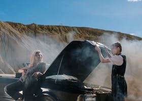 Two people standing by a smoking car with an open hood in a desert landscape.