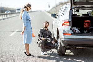 Man changing a flat tire on a silver SUV on a road. Woman watches nearby, both beside open trunk.