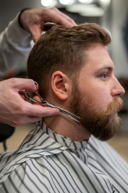 Barber trimming a man's beard with scissors in a striped cape