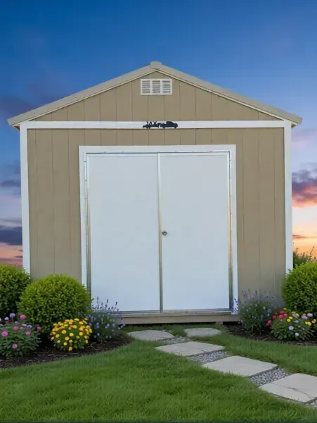 Tan garden shed with white double doors, flower beds, and a stone path at sunset