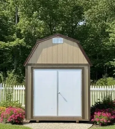 Small beige storage shed with white double doors in a flowered yard, framed by a white picket fence and trees