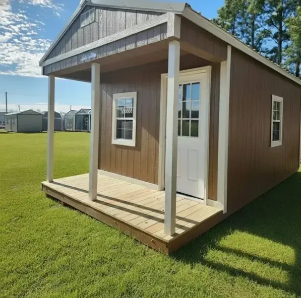 Small brown cabin with white trim and porch on a grassy lawn under a blue sky