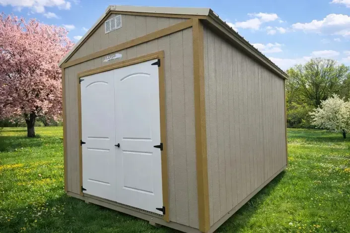 Tan portable shed with white double doors on grass, with pink flowering trees in the background