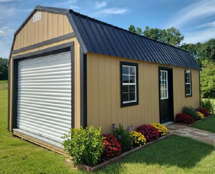 Small tan storage shed with black roof, white roll-up door, and flower beds beside a lawn.