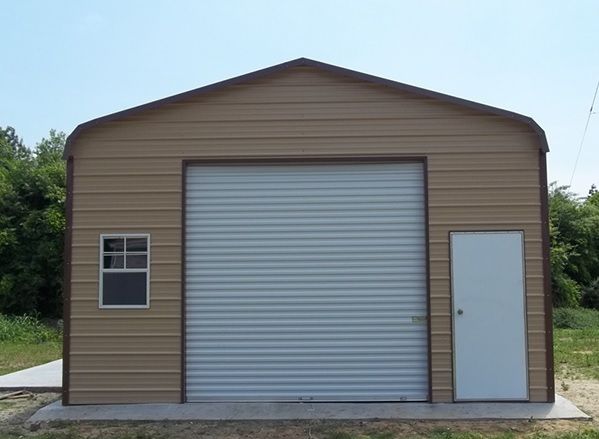 Small tan metal shed with a large roll-up door and side entry door on a gravel pad.