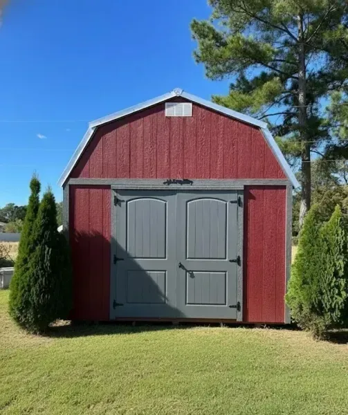 Red barn-style shed with gray double doors, framed by tall evergreens on a grassy lawn under a blue sky