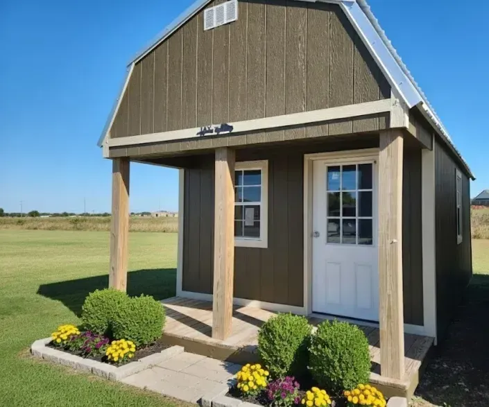 Small brown shed with white door and porch, set in a grassy field with flowers in front