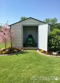 Small garden shed with open white doors, a green riding mower inside, and flower beds under a blue sky.