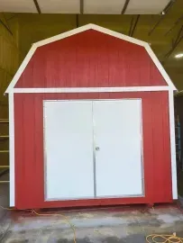 Red barn-style shed with white double doors and trim, shown indoors on a concrete floor.
