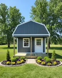 Small gray garden shed with white trim, front porch, and flower beds in a sunny green yard