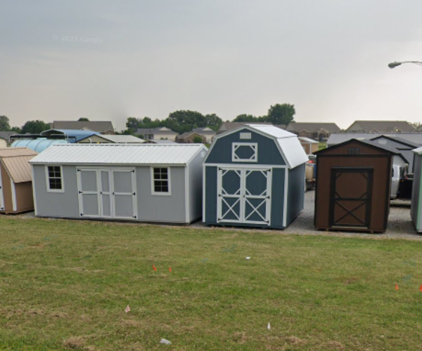 Three small backyard sheds in gray, blue, and brown on a grassy field under an overcast sky.