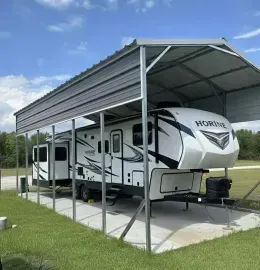 White travel trailer parked under a metal carport on a concrete pad beside a grassy field.