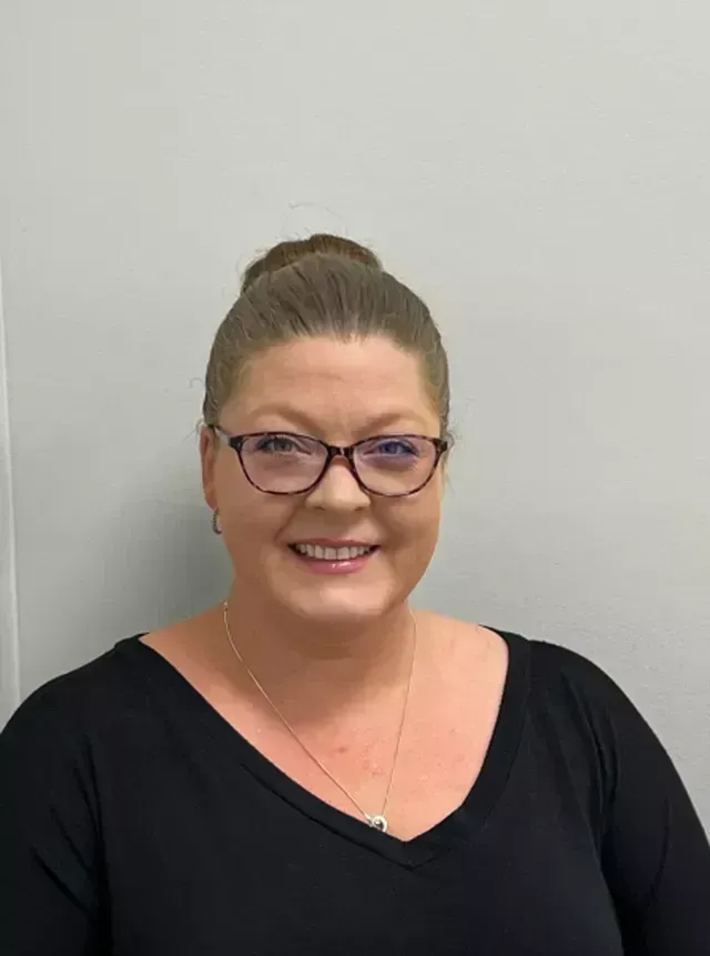 Woman with glasses and bun smiling at the camera, wearing a black top against a gray wall.