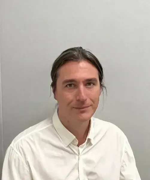 Man with brown hair pulled back, wearing a white shirt, against a gray wall, smiling slightly.