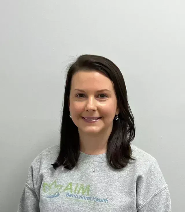 Woman wearing a gray sweatshirt smiling, in front of a gray wall. Sweatshirt has a logo that says 