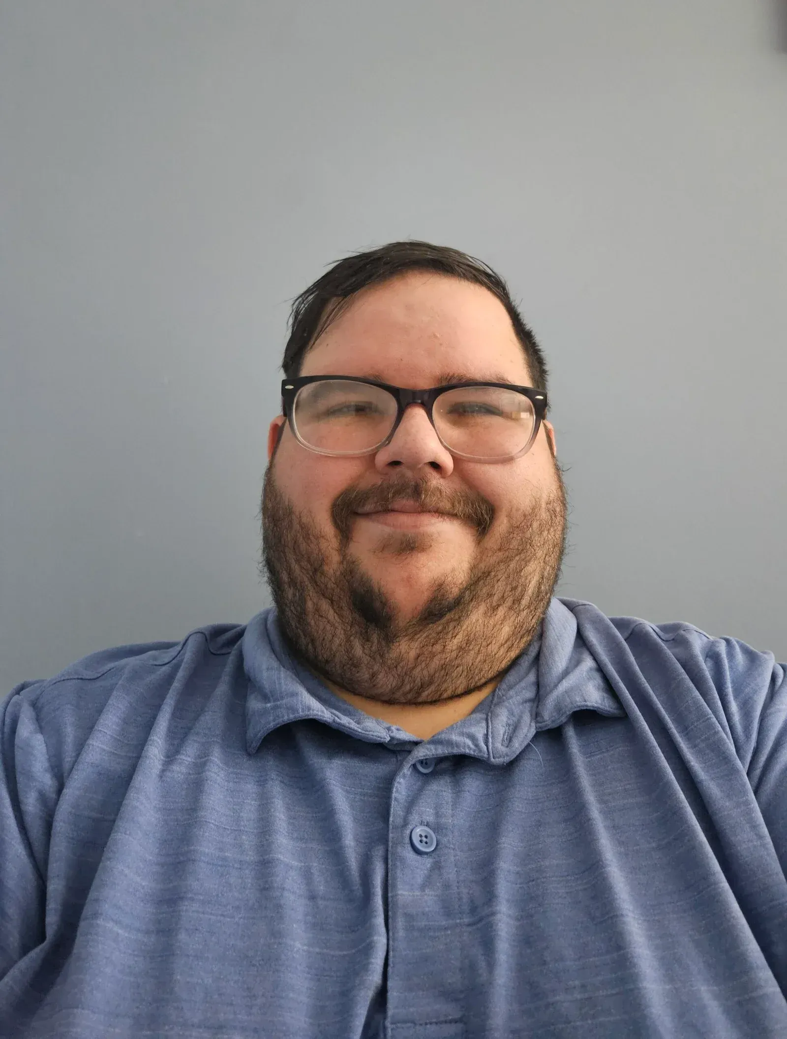 Man with glasses and a beard wearing a blue polo shirt, smiling, against a gray wall.