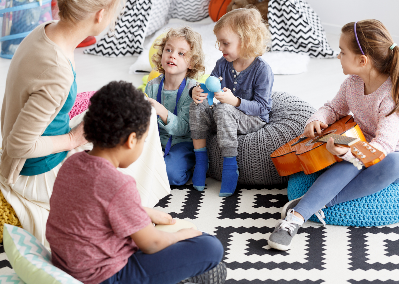 Children and teacher sit in a circle on a patterned rug, reading and holding a ukulele, listening to the teacher.