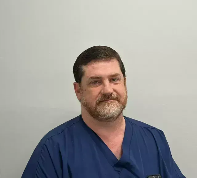 Man with beard, wearing blue scrubs, looking at the camera. Neutral background.
