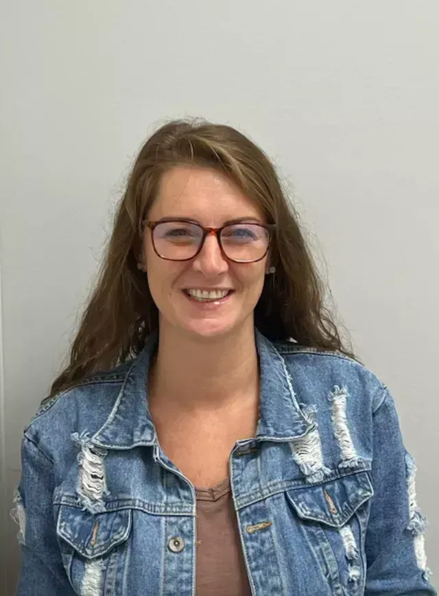 Woman with long brown hair wearing glasses and a denim jacket smiling.