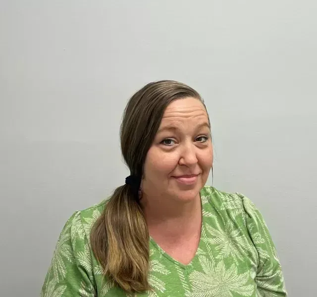 Woman with brown hair in a low ponytail smiles, wearing a green patterned shirt, against a gray background.