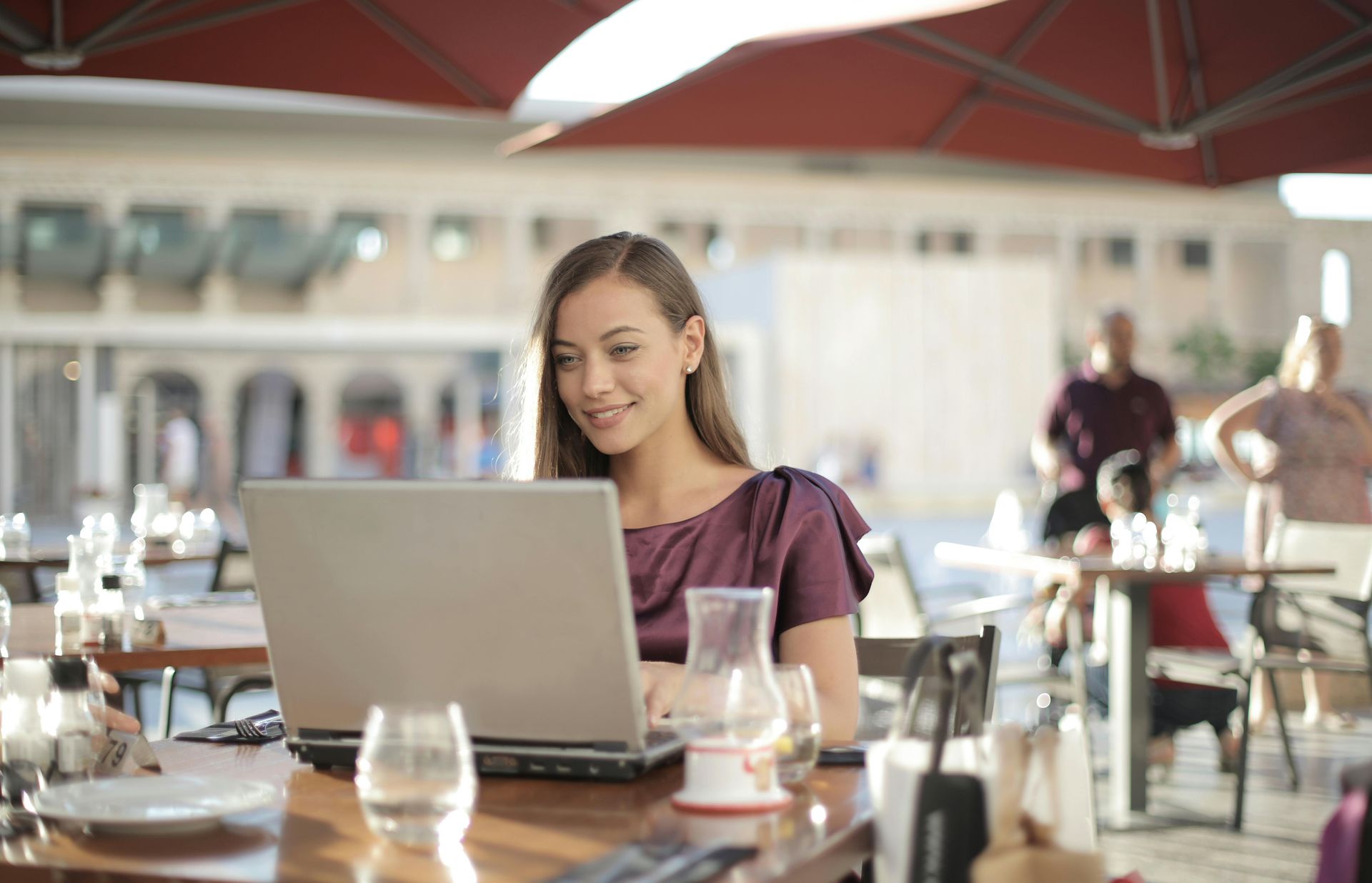 Woman smiles while working on laptop at a cafe table, outdoor setting.